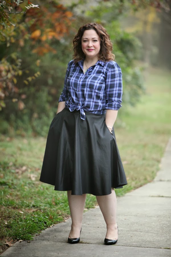 Alison Gary of Wardrobe Oxygen in a black taffeta full midi skirt with a blue plaid flannel shirt tied at the waist. She has her hands in her pockets and is wearing black pumps.