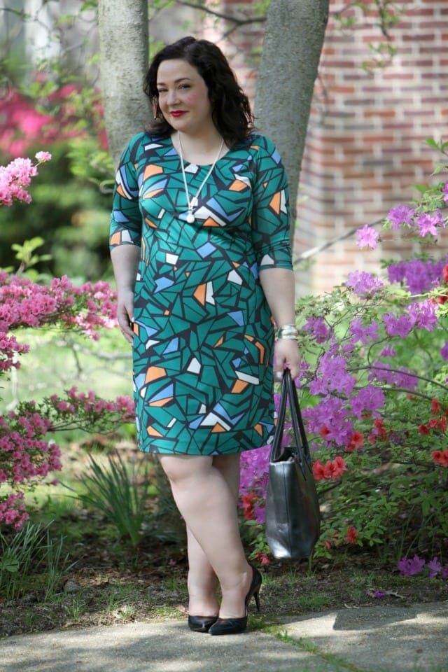 a petite size 14 woman in a green mosaic print bracelet sleeve length sheath, wearing black pumps and holding a black leather tote bag