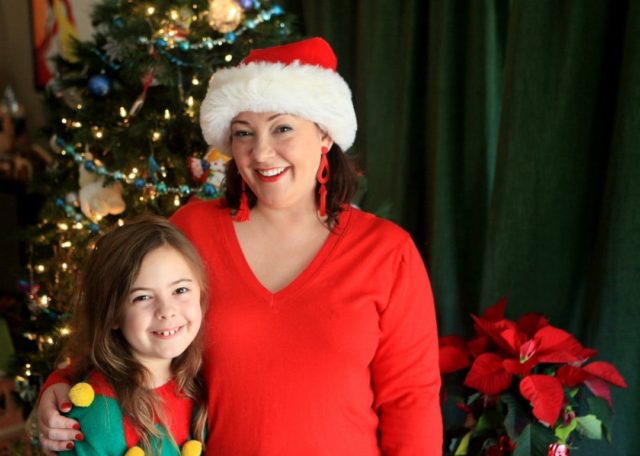 A woman in a red v-necik sweater and santa hat with her arm around a young girl in a Christmas sweater