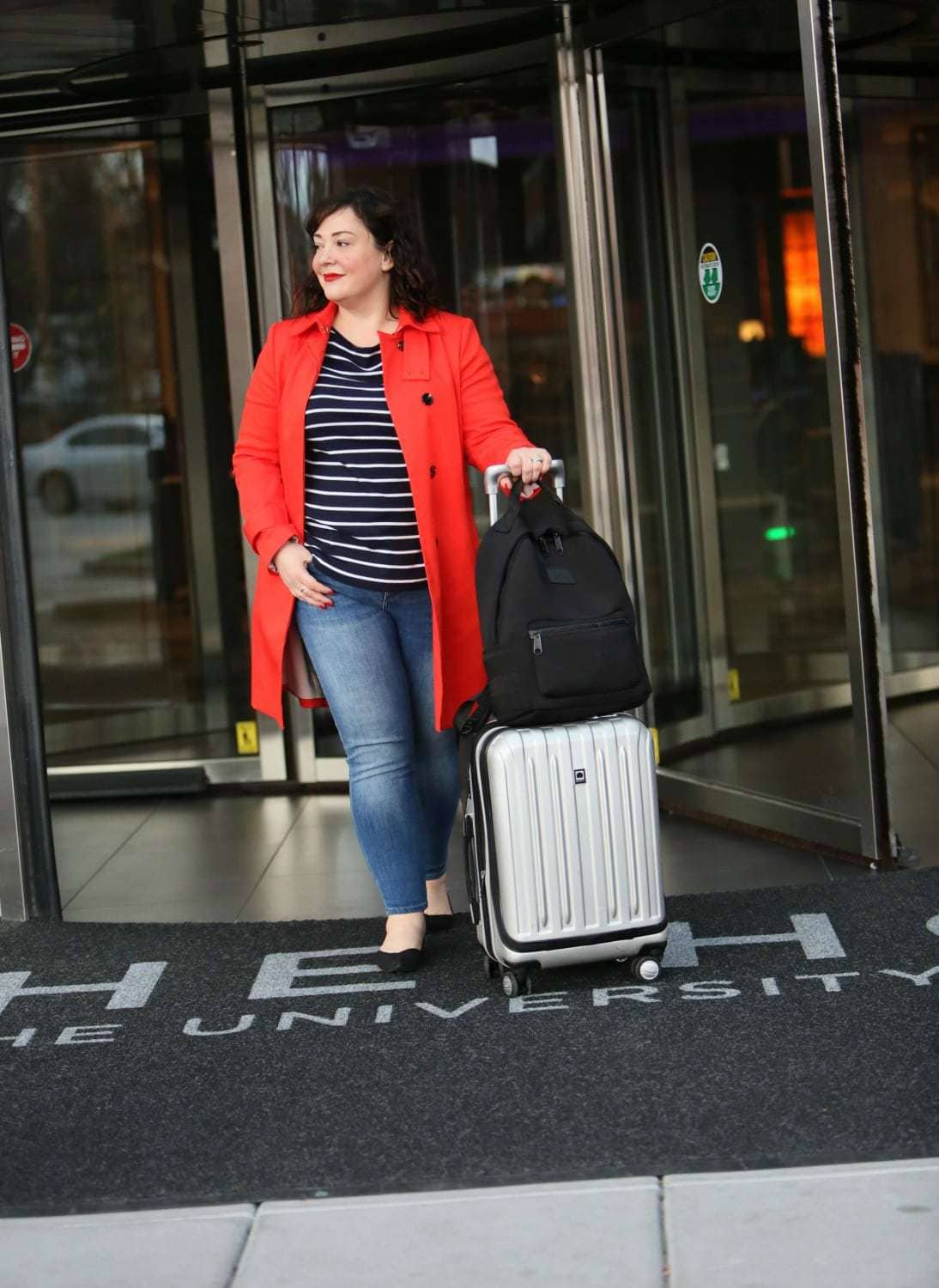 Wardrobe Oxygen in a red orange trench from Banana Republic with Talbots striped top, Banana Republic ankle jeans, Rothy's flats, Dagne Dover backpack and Delsey spinner suitcase