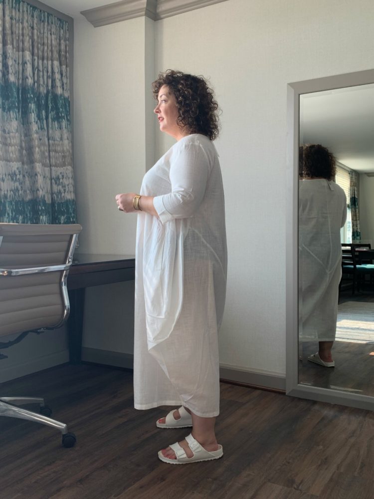 Alison Gary in an ankle-length thin cotton dress standing in profile looking out of a window for a DUBGEE by Whoopi review of the clothing line