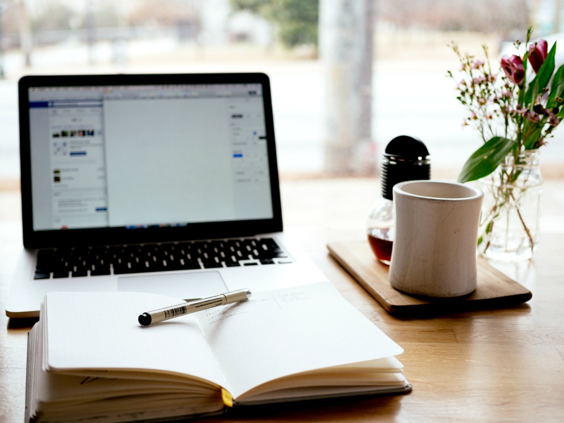 Open notebook with pen in front of open laptop with cup of coffee all on a desk in front of an open window