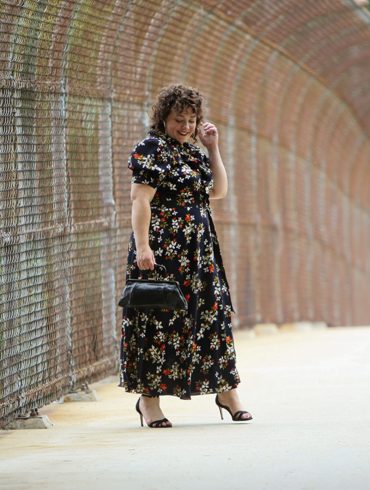 Woman in a navy floral midi length wrap dress holding a black leather vintage frame purse walking and looking at the ground