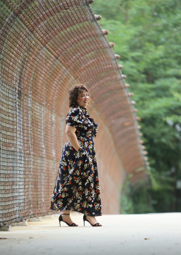 woman in a navy floral ankle length wrap dress with her hands in her pockets walking to the right and smiling at the camera