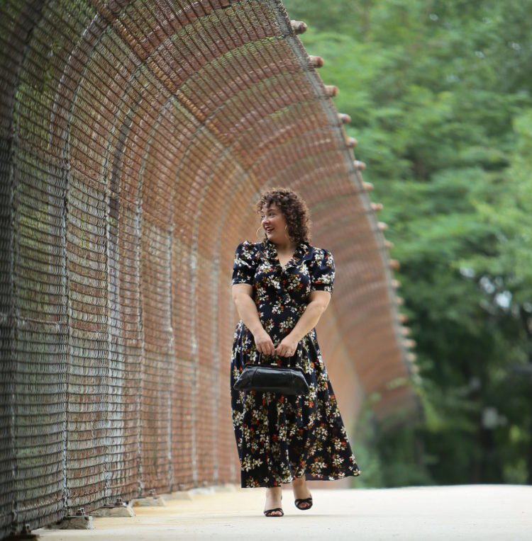 Woman in a navy floral midi dress holding a black leather frame purse in front of her looking off in the distance