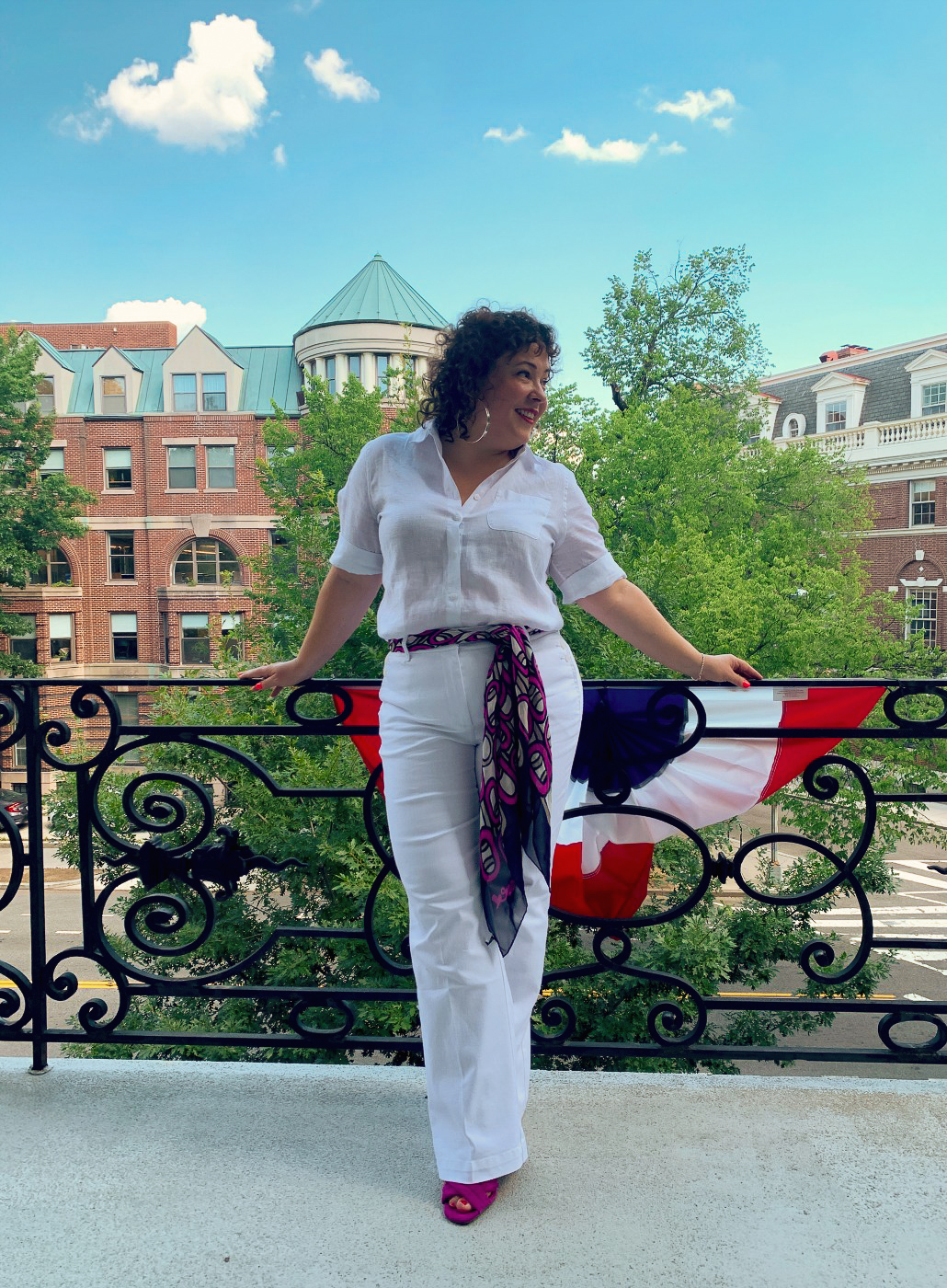 woman in white linen shirt tucked into white high rise wide leg denim trousers with a navy and magenta chiffon scarf tied around her waist as a belt. She is standing on a balcony overlooking Dupont Circle in DC and smiling.