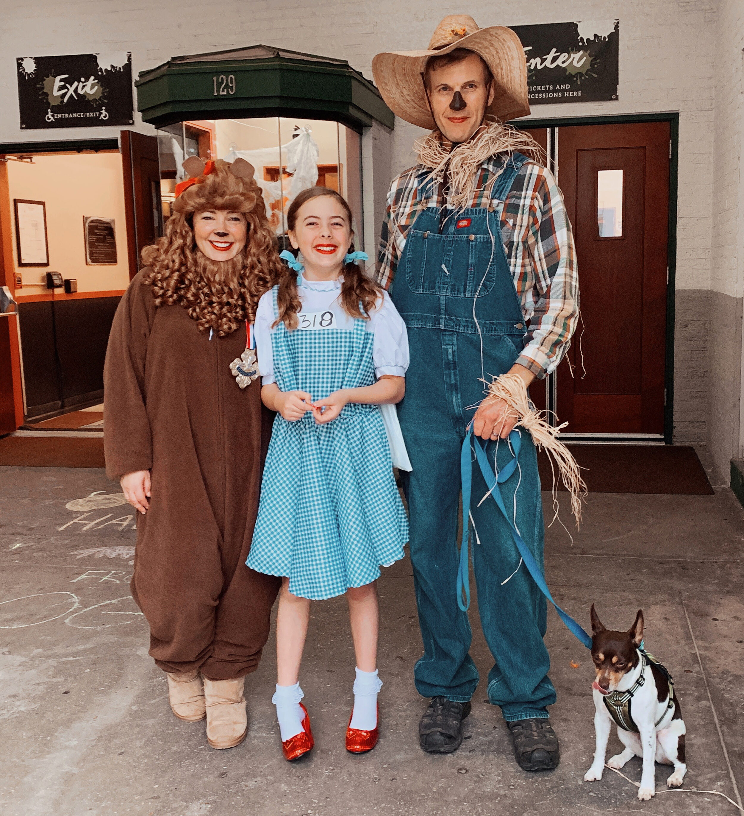 a woman dressed as the cowardly lion, young girl dressed as Dorothy Gale, man dressed as a scarecrow holding onto the leash of a small dog