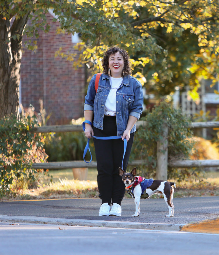 woman in a denim jacket and black knit joggers crossing the street while walking a dog