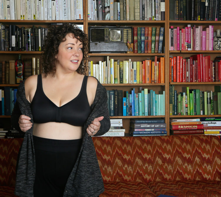 large-busted woman over 40 in a black wireless bra and black knit pants standing in front of a colorful bookcase