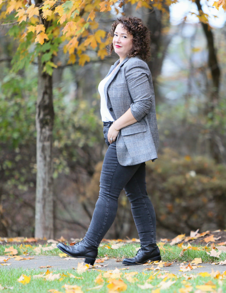 A petite curvy woman in a gray plaid oversized blazer, white v-neck t-shirt tucked into gray skinny jeans, and black lace-up boots. She is walking down a sidewalk.