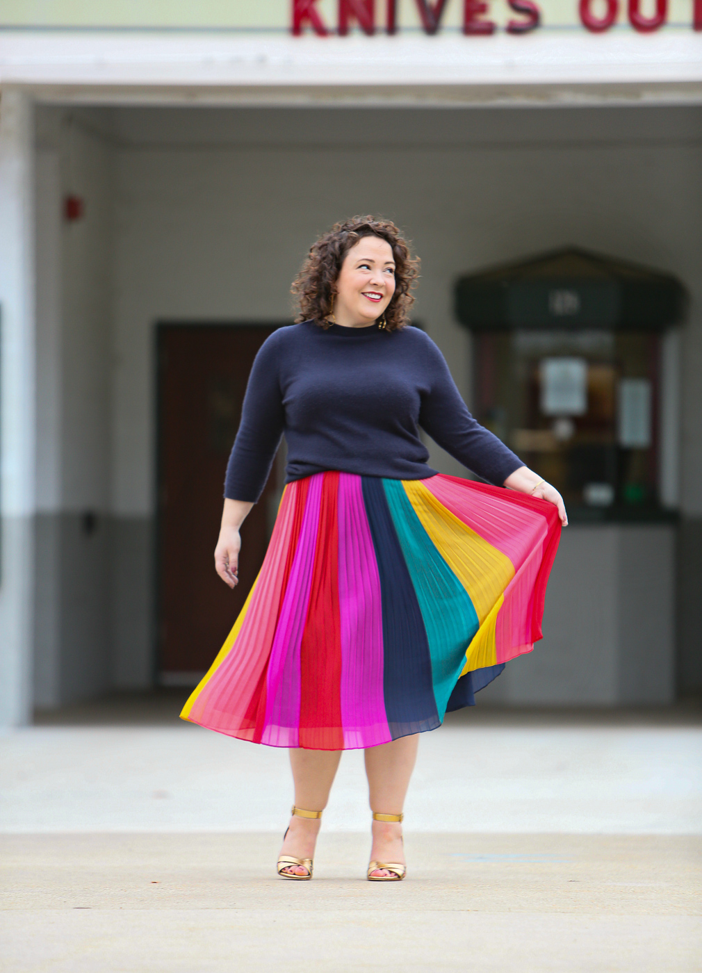 woman in a rainbow striped skirt and navy sweater standing in front of a movie theater