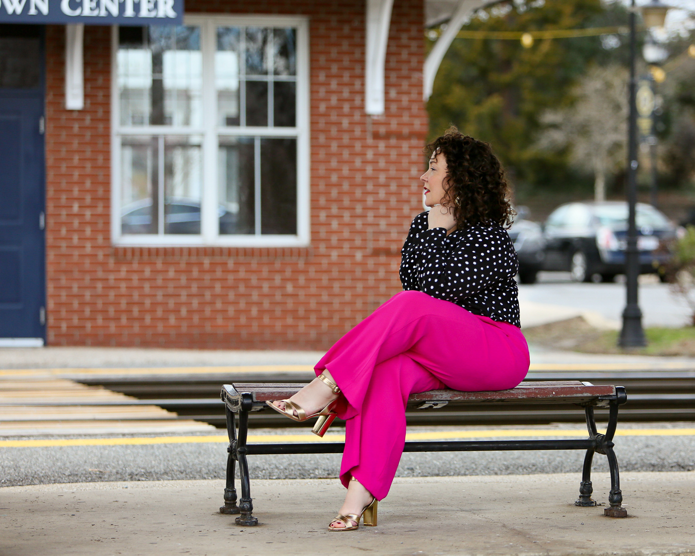 Woman in a polka dot blouse and hot pink pants with gold sandals sitting on a bench looking off into the distance