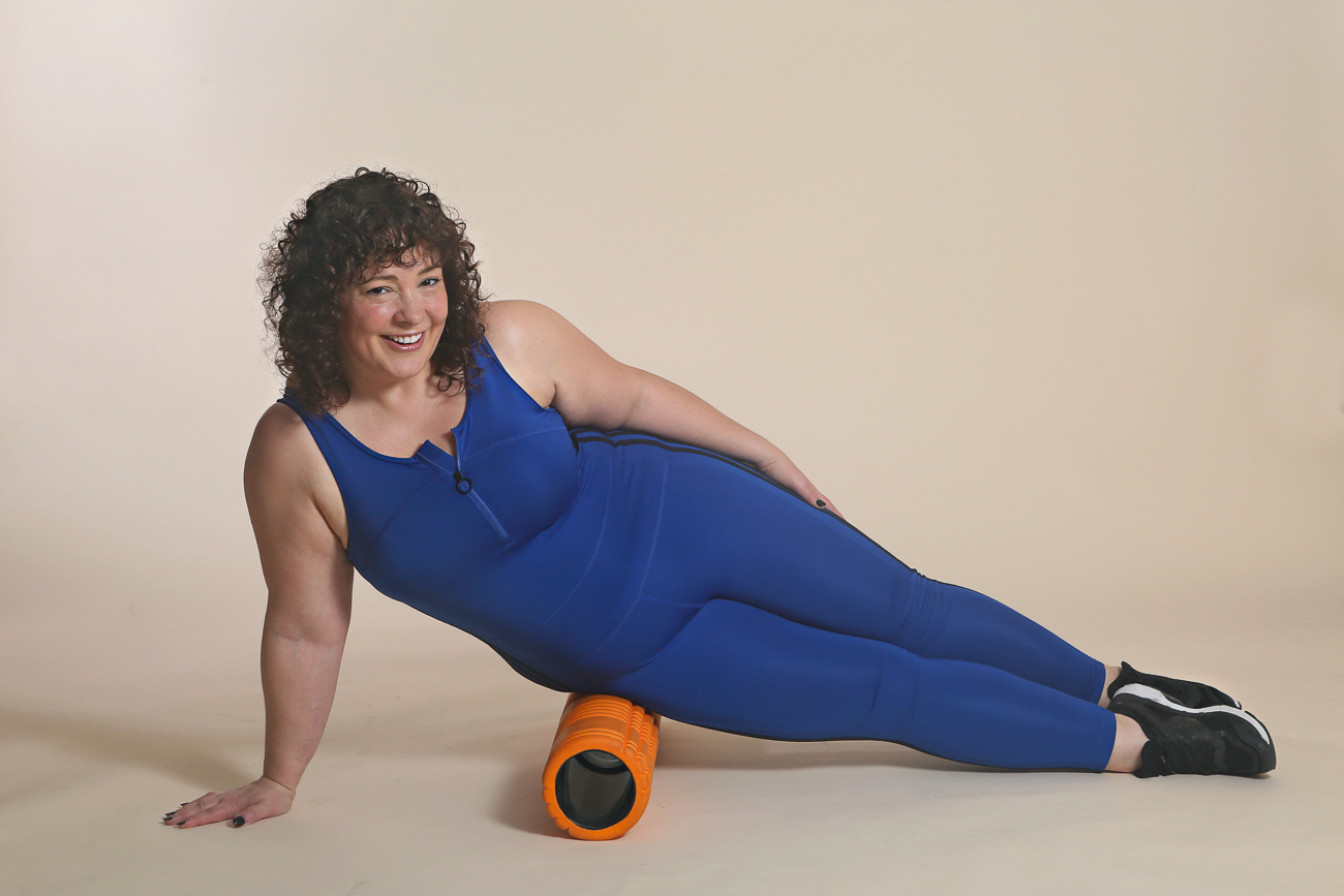 woman in blue bodysuit massaging her hip on a foam roller