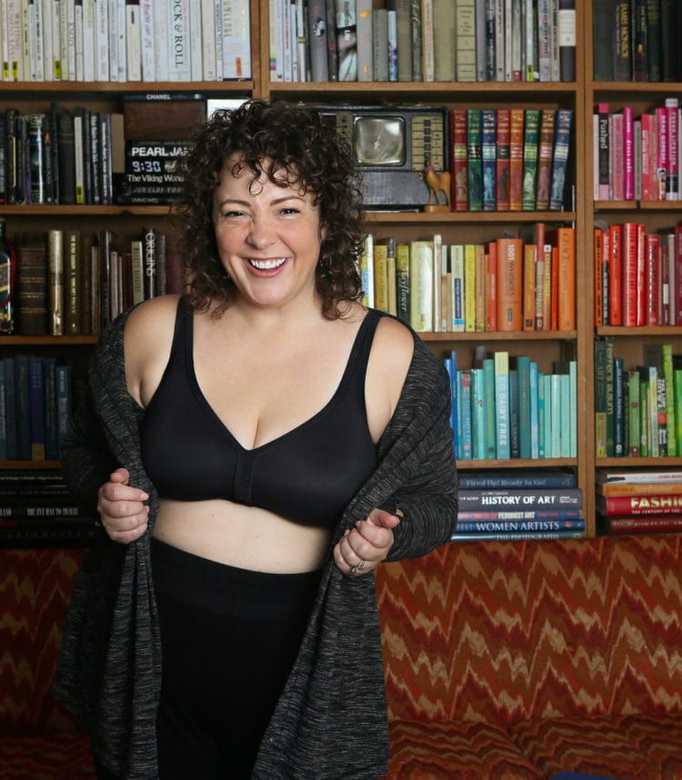 woman with curly brown hair in a living room in front of color-coded bookcase. She is wearing a black wireless bra and black pants and a heather gray knit cardigan and is smiling at the camera