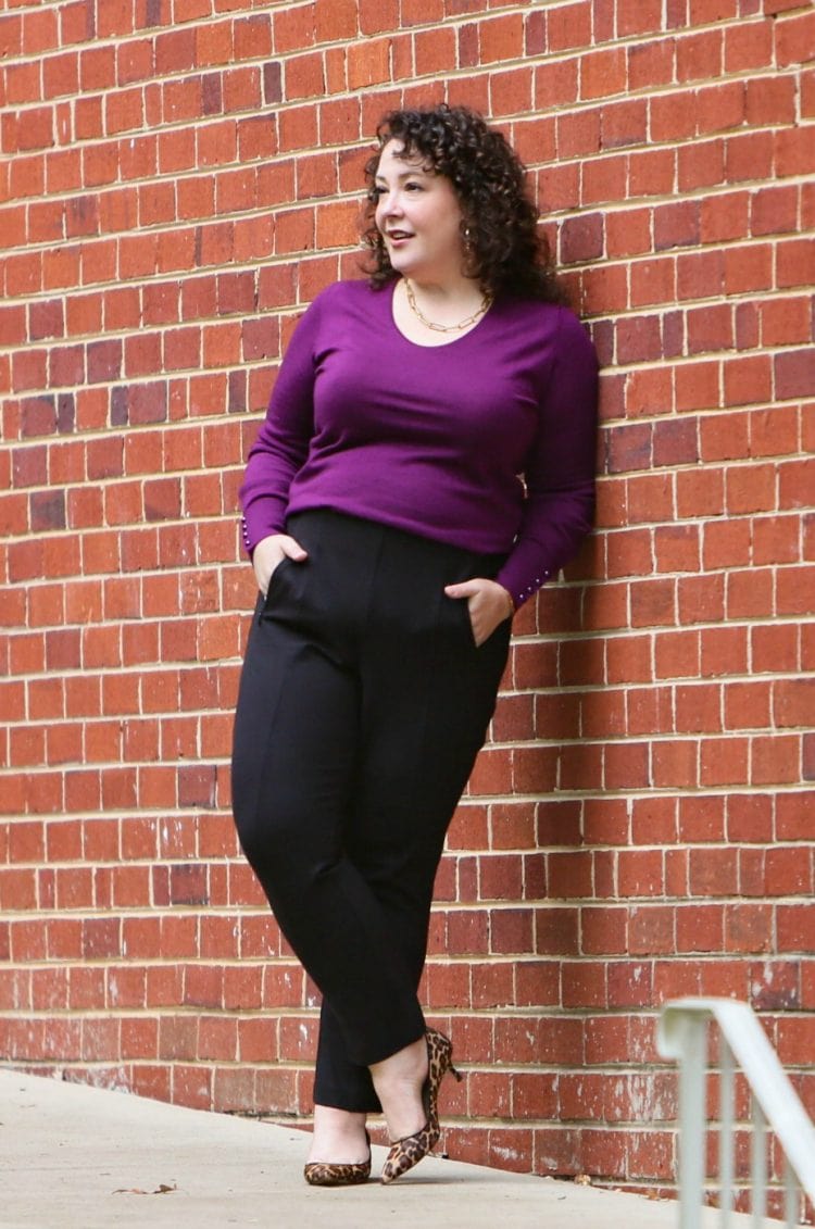 Woman in black ponte work pants and a purple merino v-neck sweater leaning against a brick wall