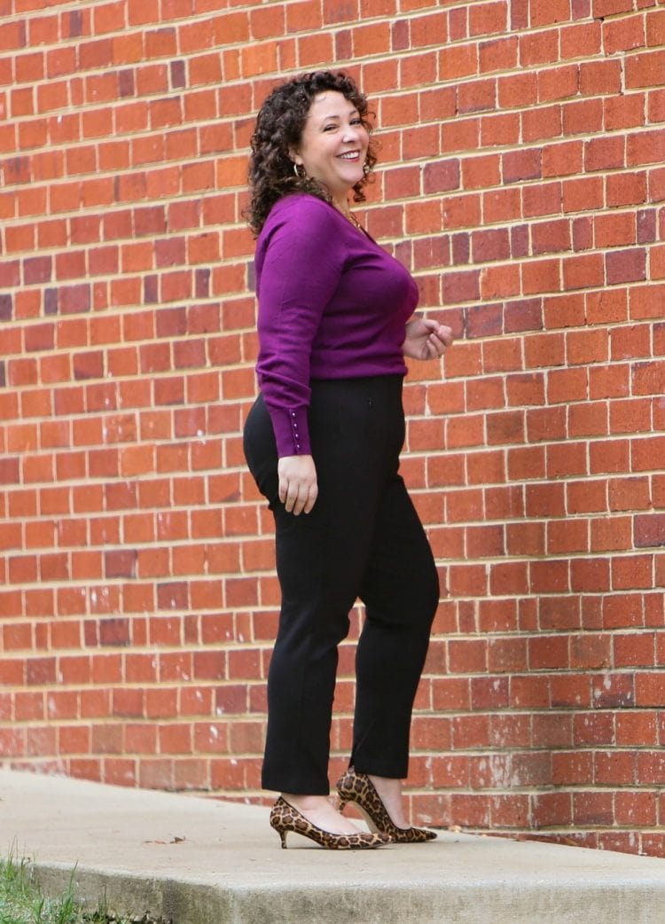 woman in black ponte ankle pants and a purple sweater smiling in front of a brick wall