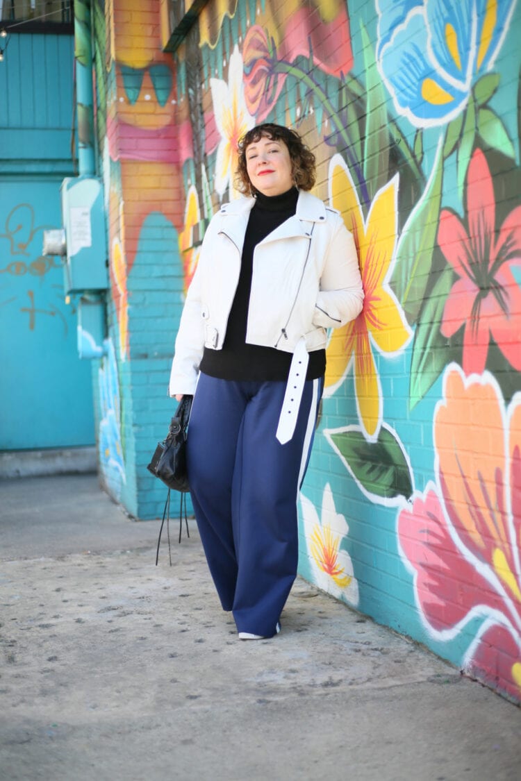 Alison leaning against a brick wall painted with a mural of tropical flowers. She is wearing a white leather moto jacket over a black turtleneck sweater with navy ponte wide leg pants and white sneakers. One hand is in her pocket and the other is holding a black leather Balenciaga City Bag.