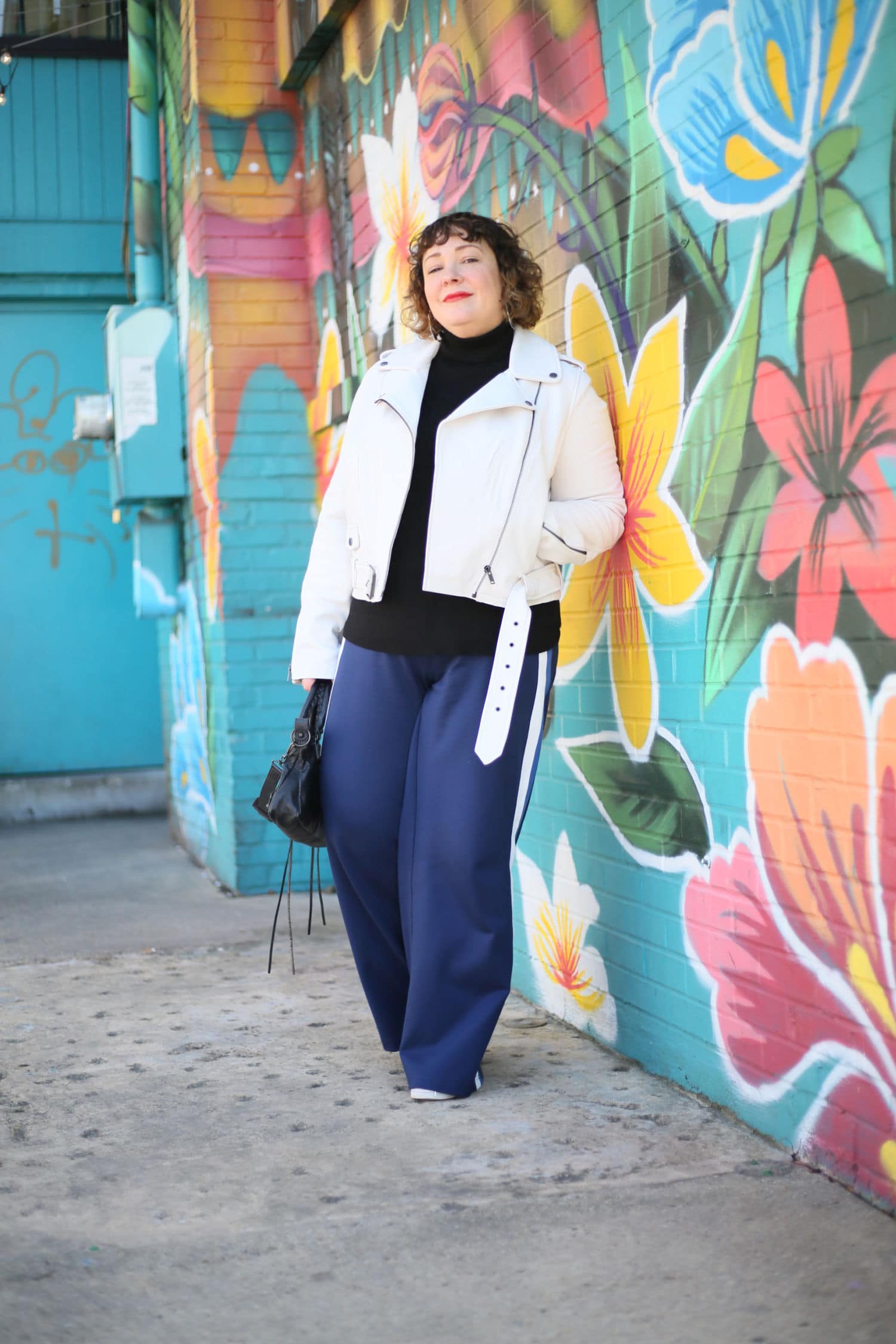 Alison leaning against a brick wall painted with a mural of tropical flowers. She is wearing a white leather moto jacket over a black turtleneck sweater with navy ponte wide leg pants and white sneakers. One hand is in her pocket and the other is holding a black leather Balenciaga City Bag.