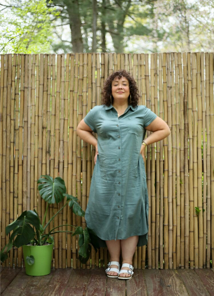 Alison wearing a green linen cap sleeve shirt dress that is midi length. She's wearing it with white Birkenstock Arizona sandals and has her hands on her hips smiling at the camera.