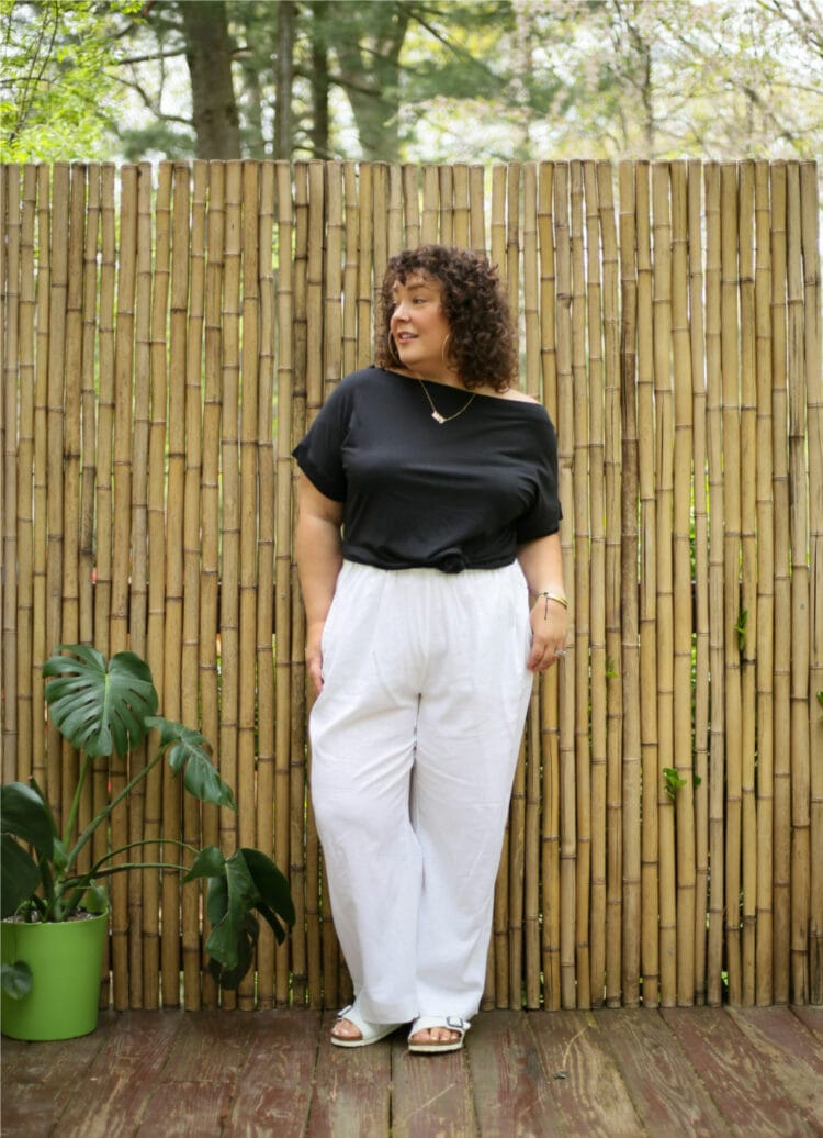 Alison in a black off the shoulder t-shirt knitted at the waist and white linen wide leg pants. She is standing in front of a bamboo wall and looking to the left.