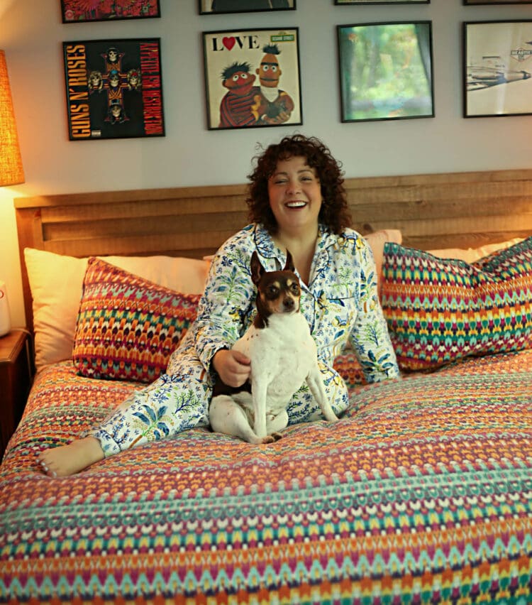 Alison wearing white and blue printed cotton full length long sleeved pajamas from Printfresh. She is sitting on her bed which is covered by a rainbow striped quilt and is cudding her small fox terrier mix dog Oscar.