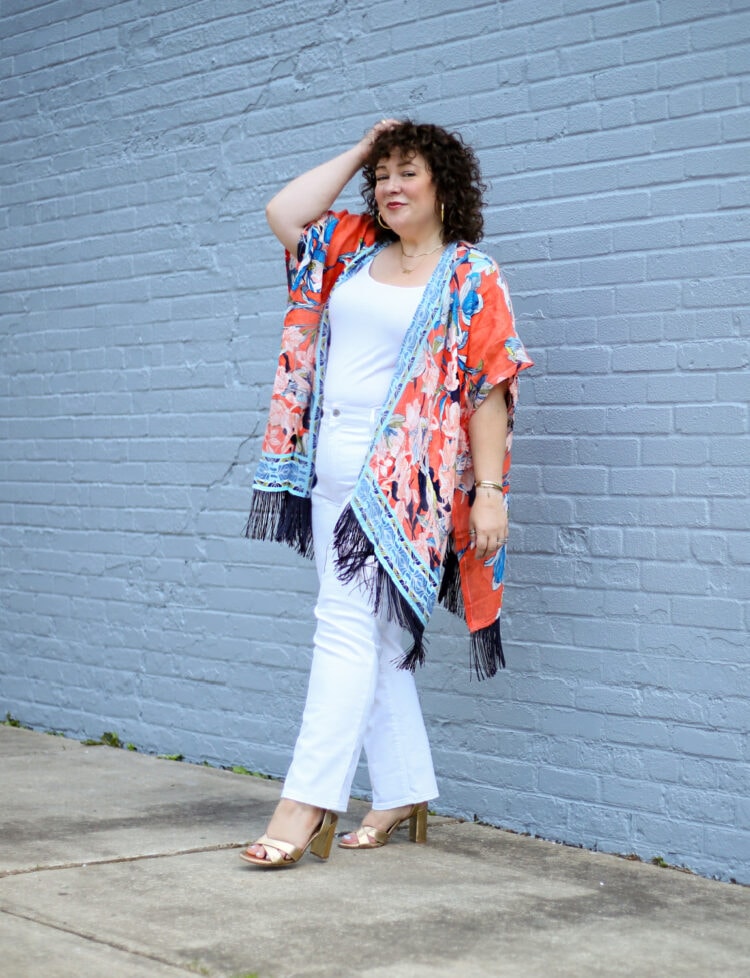 Alison of Wardrobe Oxygen wearing a linen ruana from Chico's in an orange and blue print with black tassels. She is walking away from a gray wall, her hand in her hair