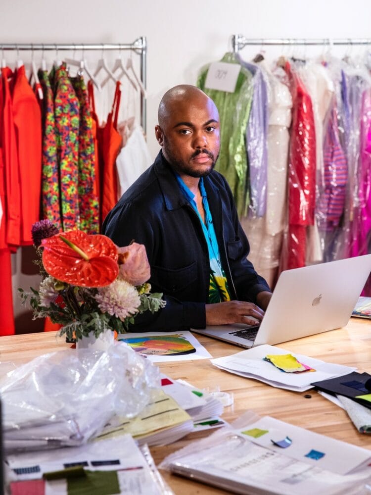 Fashion designer Christopher John Rogers sitting in his studio with racks of his colorful clothing behind him. He is wearing black jacket over a tropical print shirt. He is typing on an Apple computer and staring at the camera.