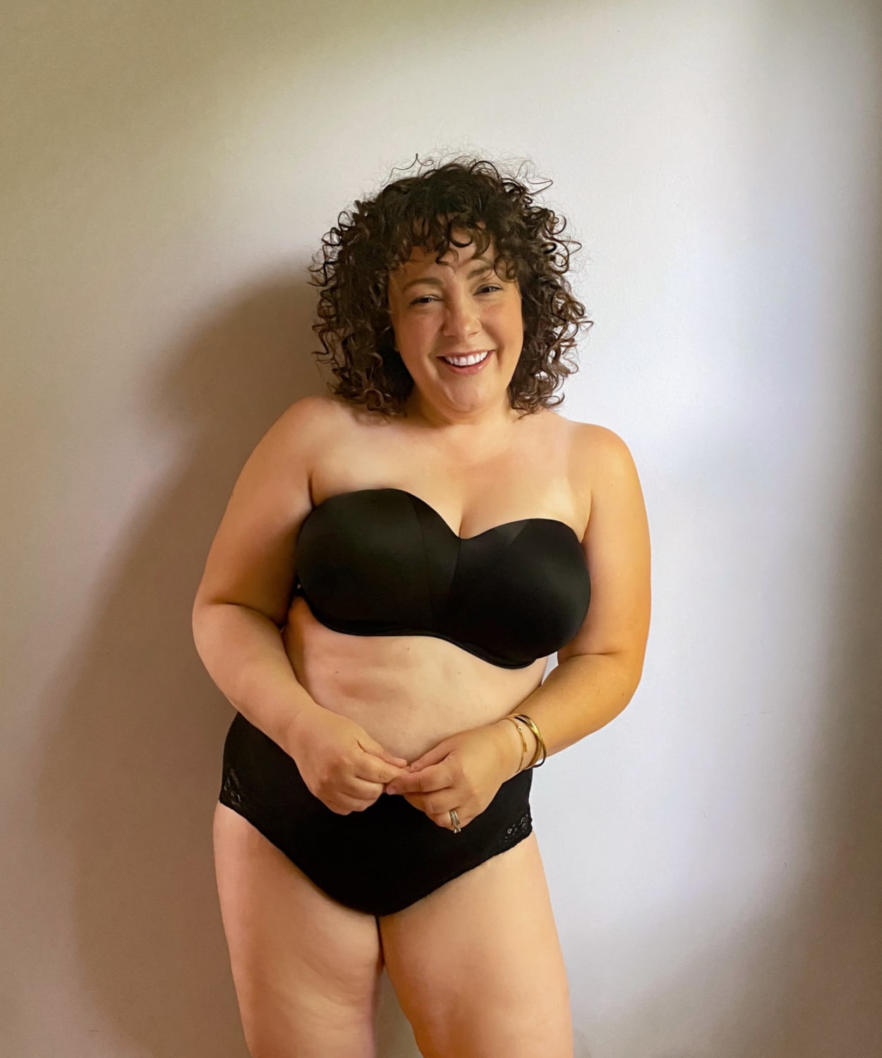 Alison wearing a black Soma Stunning Starlet strapless bra with black briefs. She is standing against a wall and smiling at the camera