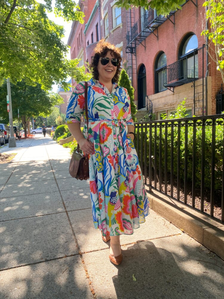 Alison walking down a sidewalk in the Dupont Circle part of Washington DC, wearing a colorful midi length Boden cotton shirtdress, sunglasses, brown leather flats and a brown leather saddle bag style purse.