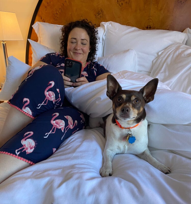 Alison Gary of Wardrobe Oxygen lying on a hotel bed wearing Soma pajamas, next to a small dog