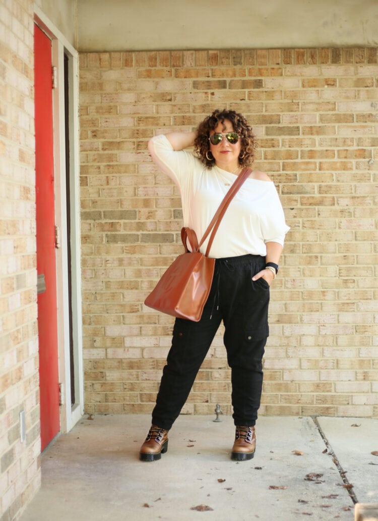 Wardrobe Oxygen in a white ALLSAINTS Rita off the shoulder long sleeved top with black jogger inspired black cargo pants from Banana Republic and a tan TELFAR medium tote worn as a crossbody. She is standing in front of a brick wall wearing sunglasses and facing the camera, one hand in her hair. 