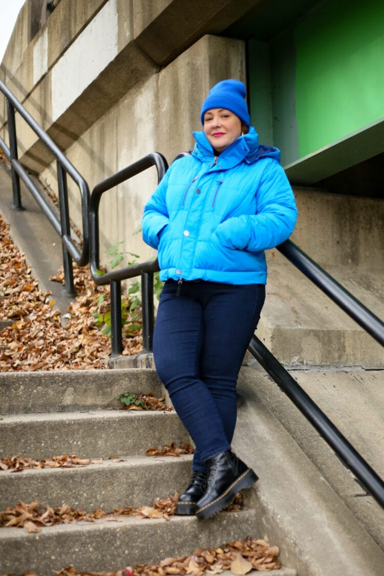 Alison wearing the short version of the Universal Standard 4-in-1 Hybrid Puffer in electric blue. She has it zipped up and her hands are in the pockets. She is wearing it with dark skinny jeans and black Doc Marten boots and a blue Universal Standard beanie on her head. Her legs are crossed and she's leaning against a railing on a concrete stairwell outside in an industrial area.