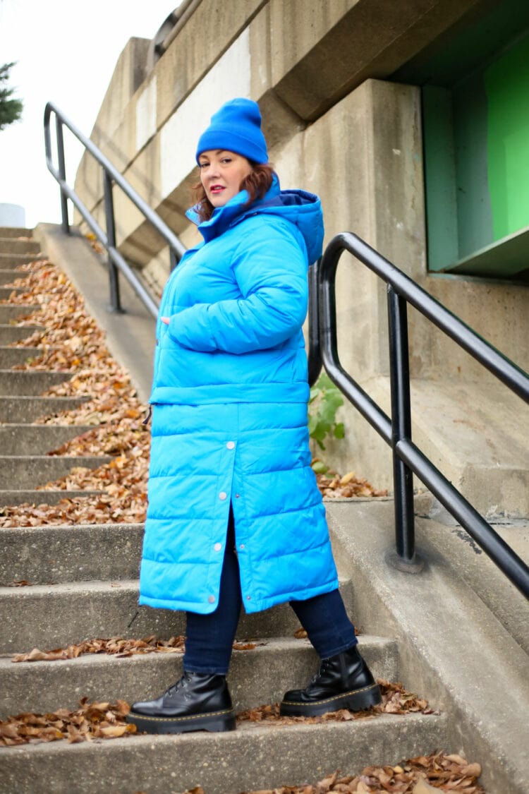 Alison walking to the left of the camera. She has the full length of the coat on, the snaps of the slits open to offer movement. She has her hands in the pockets and she is looking at the camera.