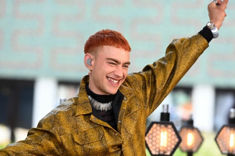 Image of Olly Alexander of Years and Years. He is in a yellow snakeskin jacket and black turtleneck with his hand in the air smiling.