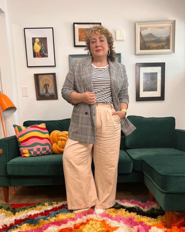 Where to Shop When You are Midsize Woman: Alison of Wardrobe Oxygen wearing a plaid M.M.LaFleur blazer with a striped Universal Standard t-shirt and wide leg pants from Madewell. She is standing in her office in front of a green velvet sectional sofa from Albany Park Home.