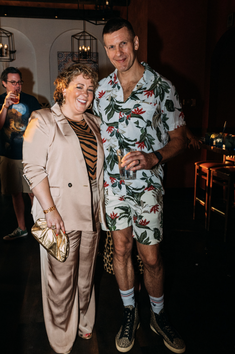 A woman in a tan satin pantsuit with a tiger print t-shirt with her arm around a tall man in a tropical print hawaiian shirt and matching shorts