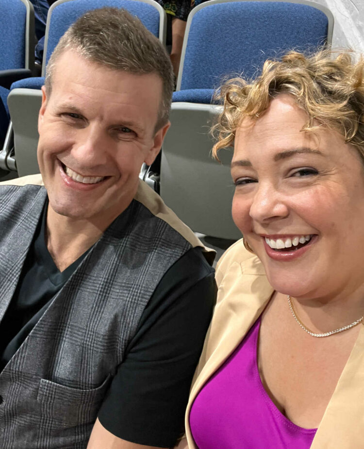 woman wearing tennis necklace sitting next to a man in a glen plaid vest and black t-shirt.