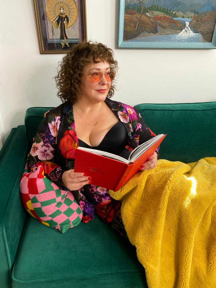 Alison of Wardrobe Oxygen in Soma Cool Nights pajamas as she lounges on a green velvet couch, covered with a gold faux fur throw, reading a book about Diana Vreeland and wearing pink tinted reading glasses from CADDIS