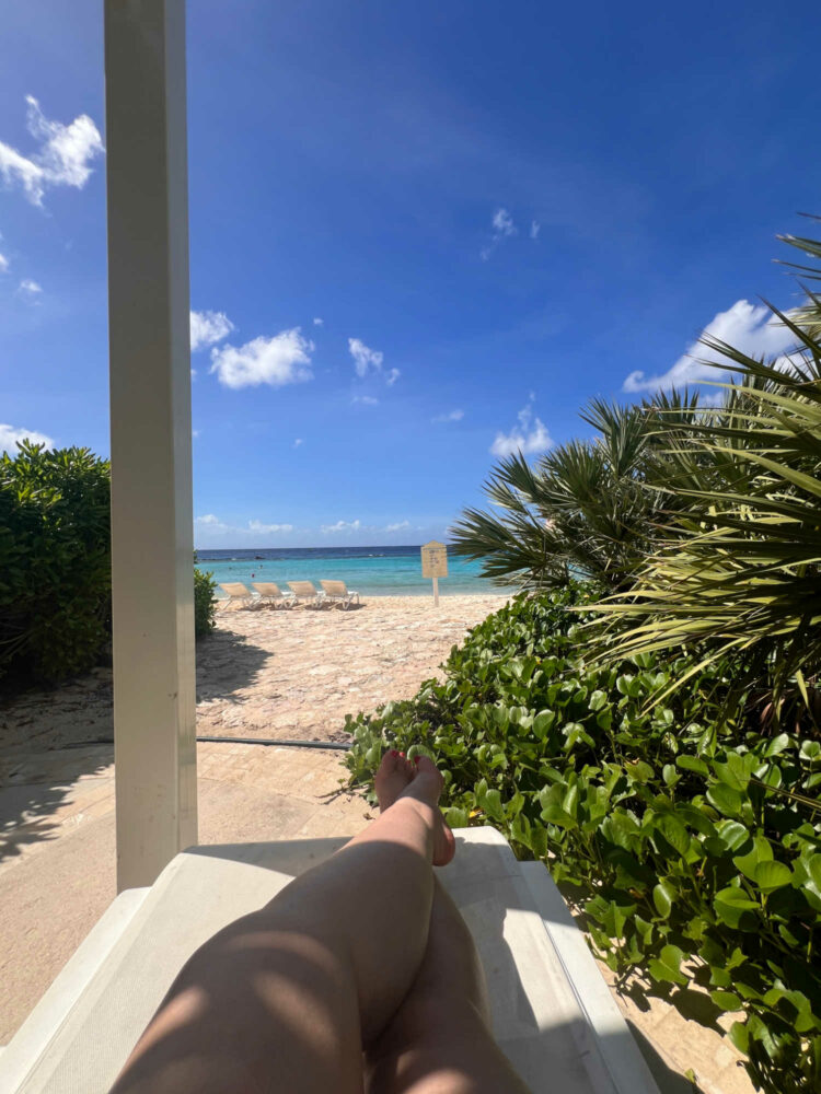 a person's legs crossed at the ankle on a lounge chair overlooking an empty beach with greenery on either side