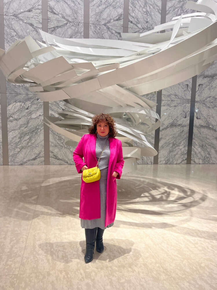 Woman in a gray Quince cashmere turtleneck and matching pleated midi skirt. Over it she is wearing a hot pink wool below knee length coat. She has a pale lime green quilted Coach crossbody over it. On her feet are black knee high boots from Ros Hommerson. She is standing in front of a modern sculpture in the lobby of an office building.