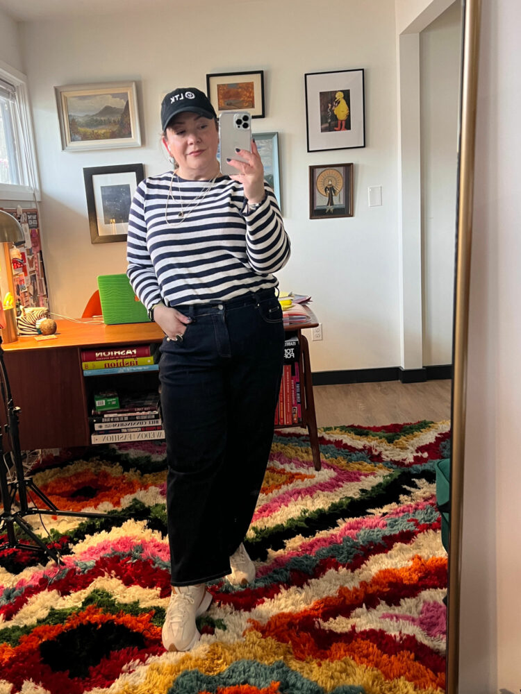 Woman in a black baseball cap, navy and white striped Breton top tucked into the dark selvedge wash of Universal Standard Bae Jeans.