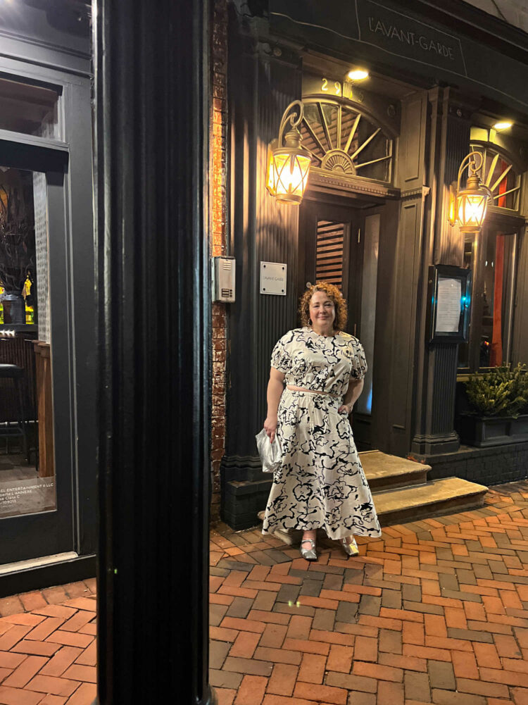 Alison Gary, a petite over-40 woman with curly brown hair is standing outside L'Avant-Garde in Georgetown. She is wearing a black and white patterned puff sleeve top and ankle length skirt. A lavender pashima is over her shoulders and she is holding a silver clutch purse.