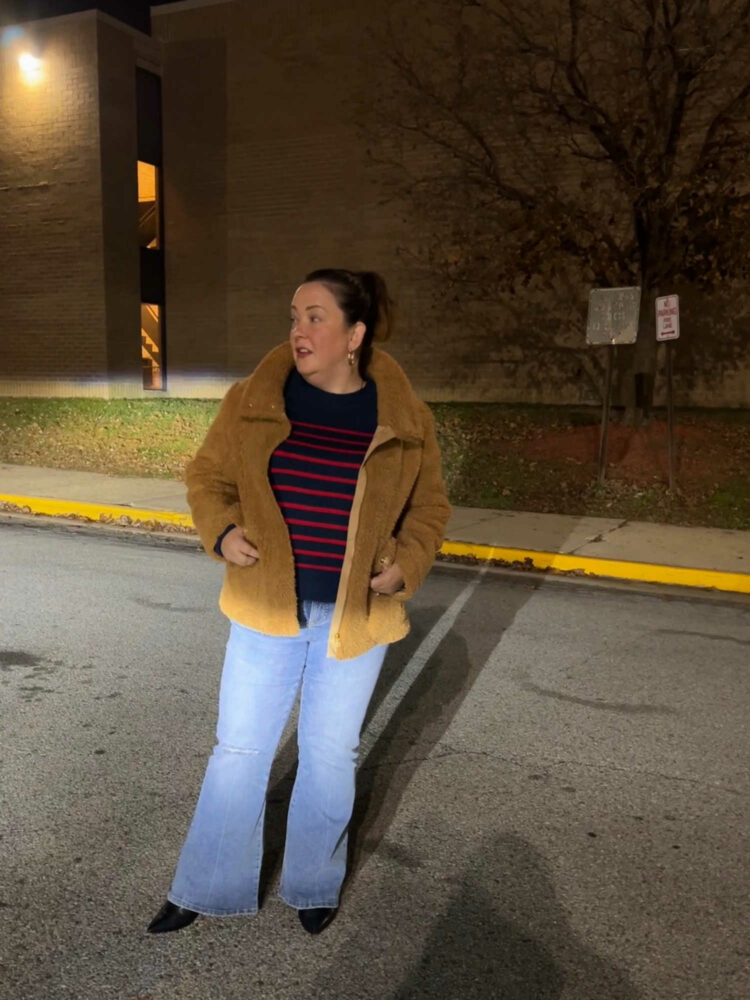 tan teddy short coat, a navy sweater with red stripes, and a faded pair of the Universal Standard Farrah flared jeans