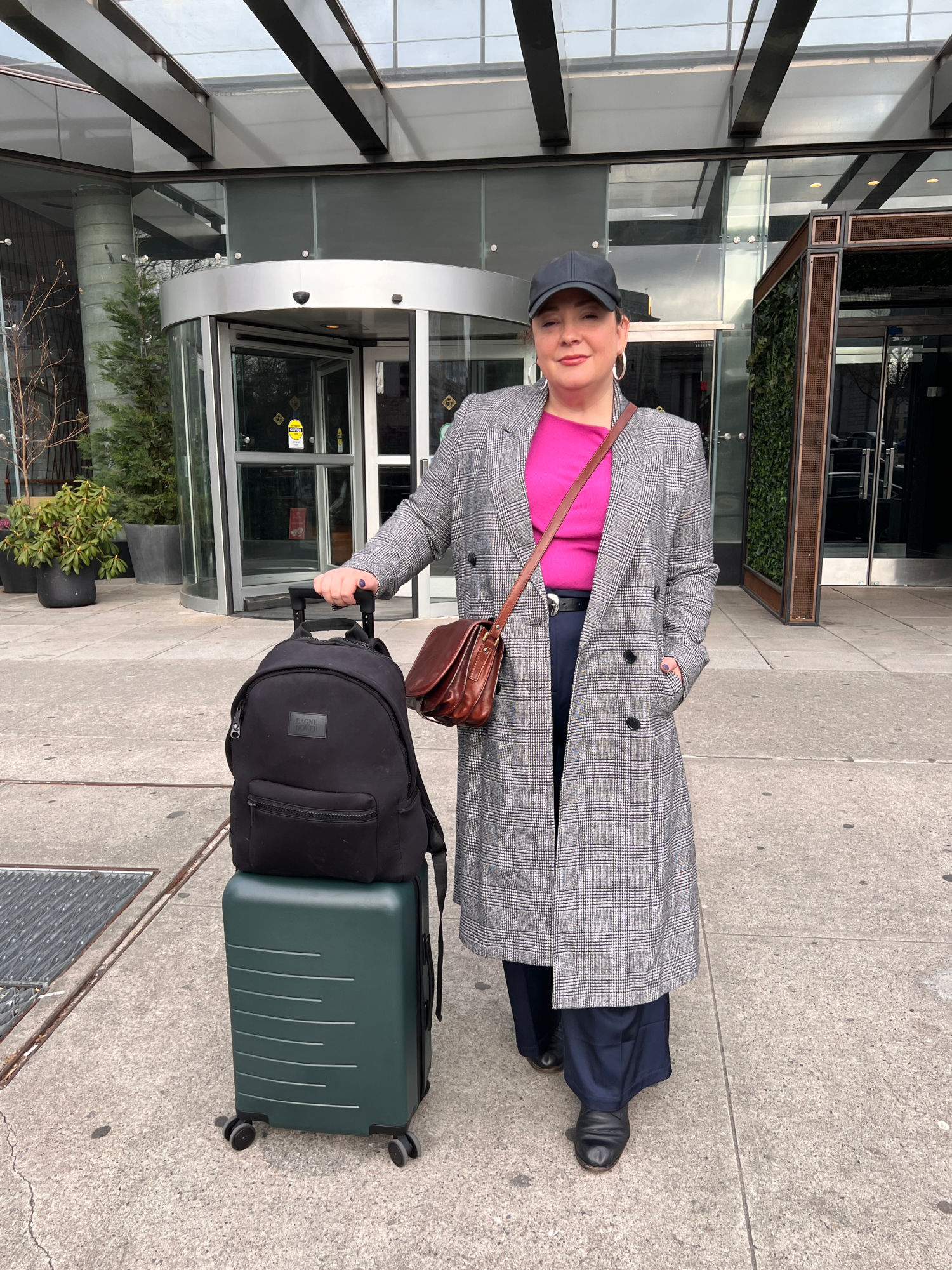 Alison Gary of Wardrobe Oxygen outside an NYC hotel holding onto a Quince rolling suitcase that has a Dagne Dover Dakota backpack on top of it.