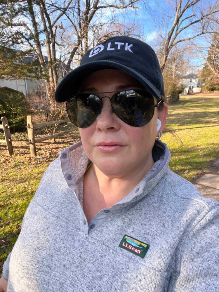 alison gary of wardrobe oxygen walking through her neighborhood listening to an audiobook. She is wearing an LLBean sweater fleece and a black baseball cap from LTK