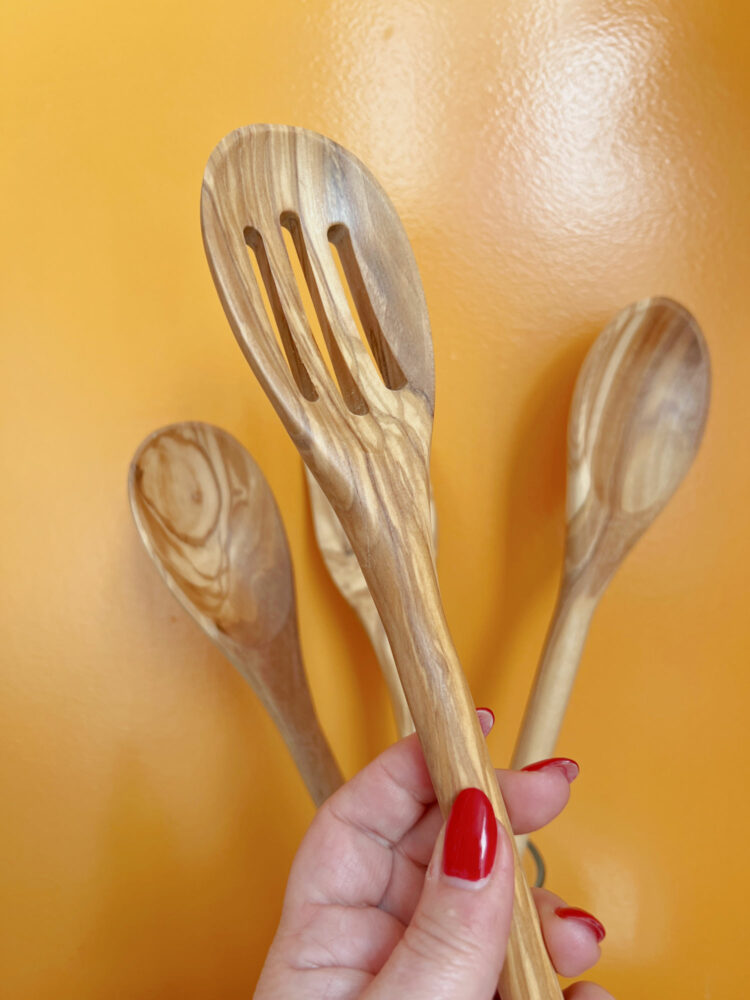 a woman's hand holding a quince olivewood slotted spoon with other wooden kitchen utensils from quince in the background